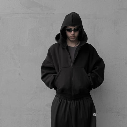 Person wearing a black hoodie and pants against a gray wall